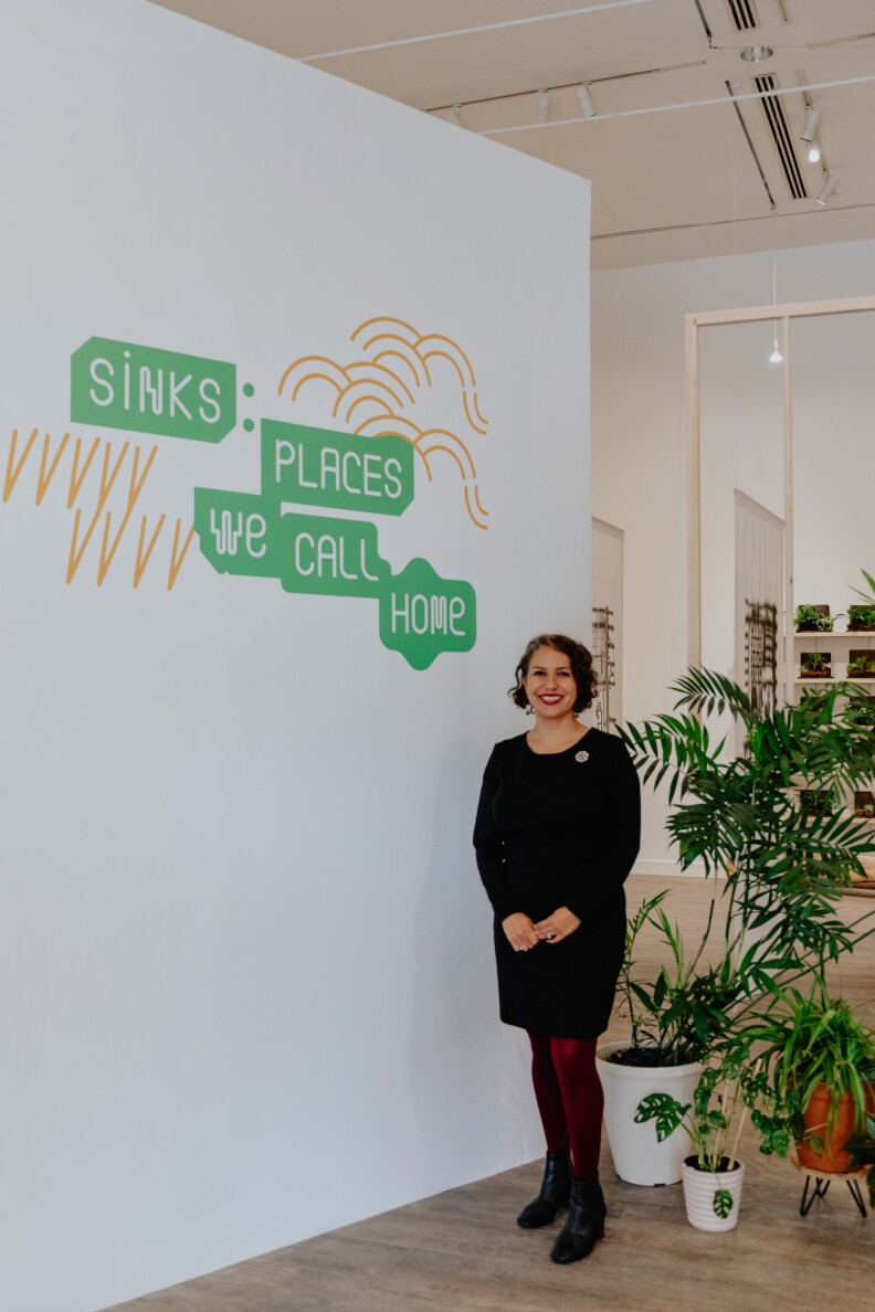 A woman with light skin tone and short medium-brown hair smiles while standing next to a sign that reads "SINKS: PLACES WE CALL HOME." Behind her there are several potted plants, of different sizes.