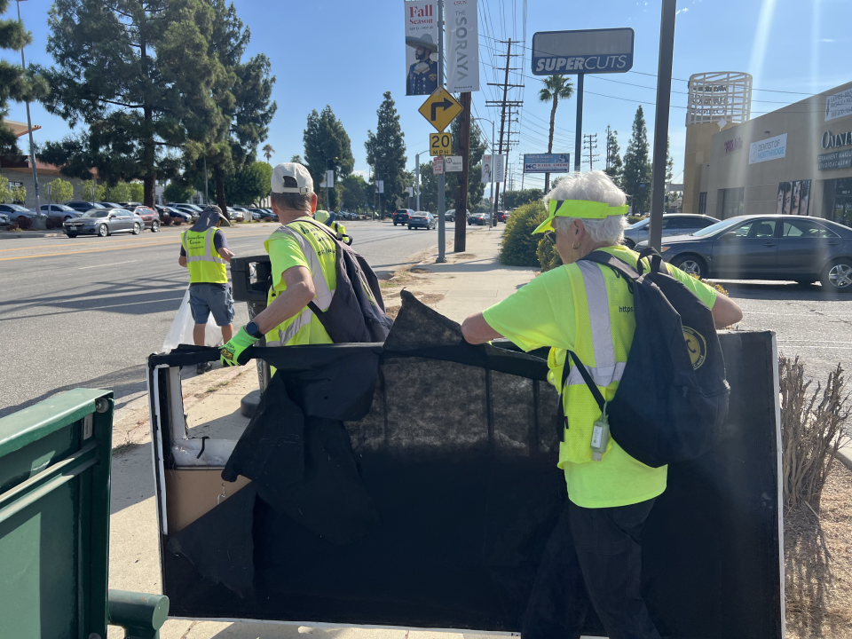 Two members of the Volunteers Cleaning Communities pick up a bed frame that was illegally dumped in a parking lot and move it to the sidewalk where the city can pick it up. The back of their heads can be seen. They are wearing yellow shirts and yellow vests.