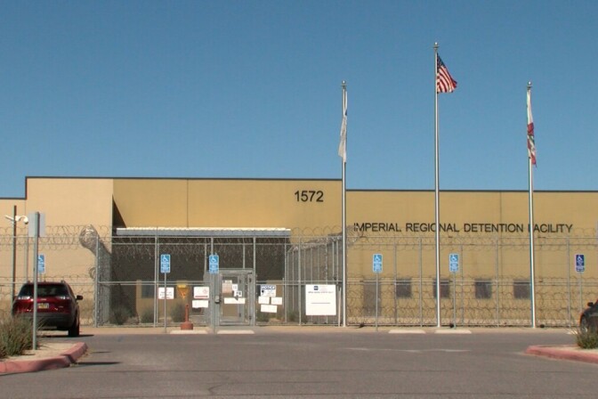 A one story, brown building behind a barbed wire fence. Three flagpoles are in front of the building. The middle flagpole is flying the American flag, the two white, unfurled flags hang on the other two poles. On the building is signage that reads, "Imperial Regional Detention Facility."