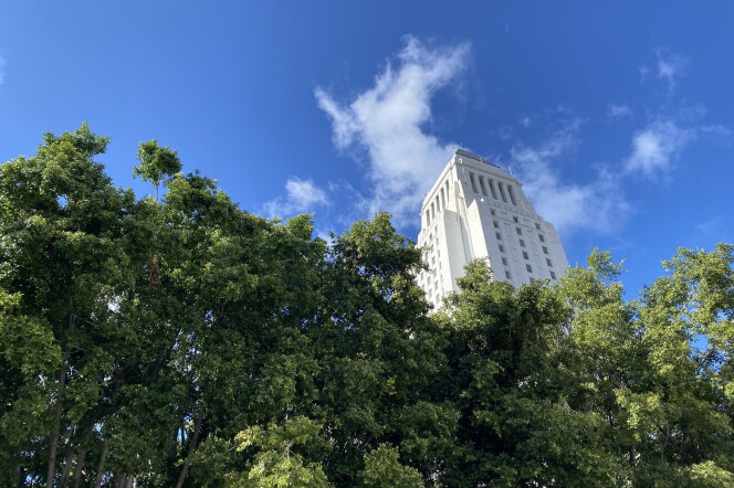 the top of city hall can be seen against a blue sky and clouds with green leafy trees in the foreground. 
