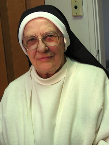 Sister Mary Raymond, wearing glasses, a white nun's habit with a black cornette, looks into the camera's lens.
