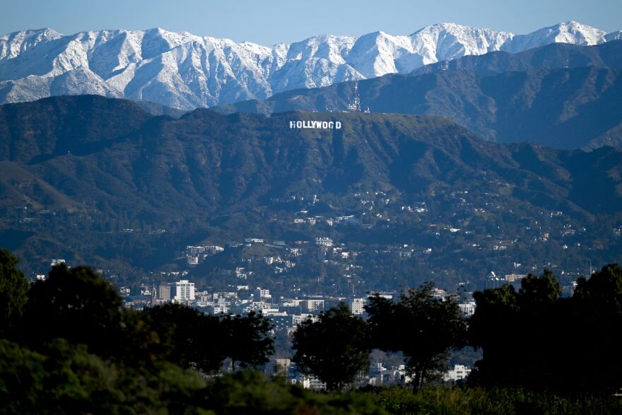 The Hollywood sign's white letters are visible on a green hillside with neighborhoods visible below and snow-capped mountains beyond.