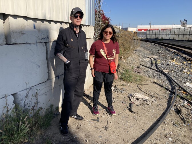 A tall man wearing black coveralls, sunglasses and a grey hat stands next to a slightly shorter woman wearing black joggers, a maroon t-shirt and sunglasses. THey're standing next to a cement block wall and near railroad tracks, with dirt and debris on the ground.