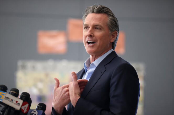 Governor Gavin Newsom is shown behind a podium with many news microphones. Newsom is wearing a navy suit with a light blue shirt. 