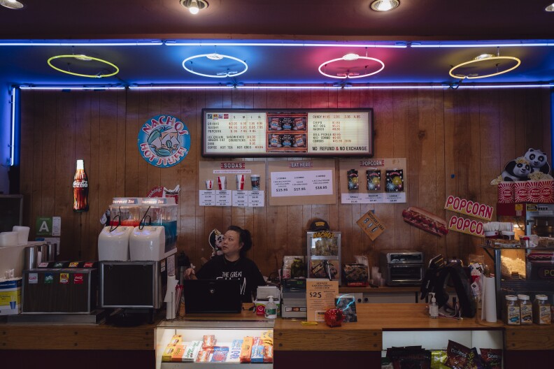 An Asian woman stands behind a counter for concessions at an older, independent movie theater.