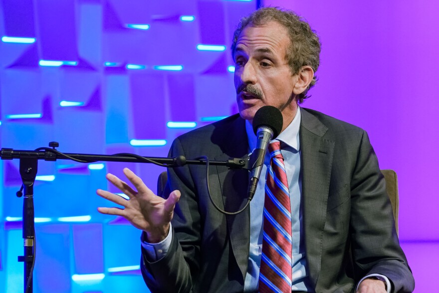 Mike Feuer wears a dark suit and a diagonal striped tie and gestures as he speaks into a mic.