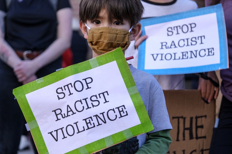 A child with a light brown face mask holds a green and white sign that says "Stop Racist Violence" in black letters. Behind the child is an adult holding a blue and white sign that says the same thing.