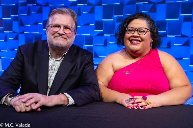 Drew Carey, a white man with a beard and glasses wearing a blue blazer and floral shirt and Yesika Salgado, a dark skinned woman with short dark hair and glasses wearing a bright pink sleeveless top and big jewel rings, looking at the camera and smiling