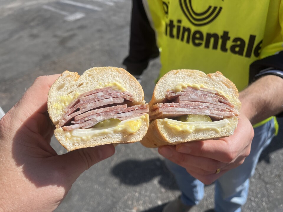 Two hands with light skin tones hold identical halves of the same sandwich next to each other. Each is made with an Italian roll with varying types of pale reddish cured meat over a slice of green pickle and white cheese underneath a layer of white mayo and yellow mustard at the bun's base. 