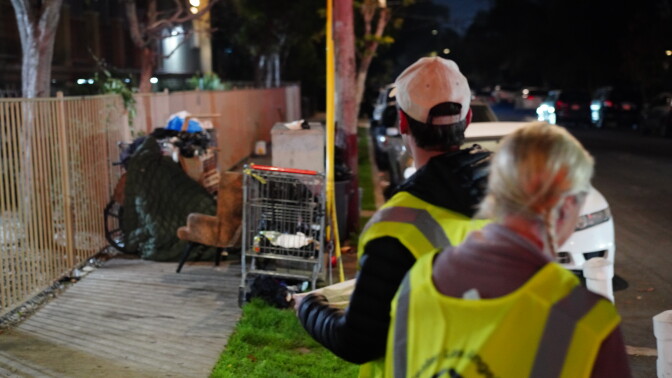 Two people wearing reflective vests stand next to a makeshift shelter on the sidewalk.
