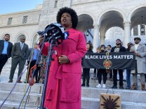BLM co-founder Patrisse Cullors speaks into several microphones outside LA city hall. A banner behind her reads "Black Lives Matter." City council members and several activists stand behind her. Cullors wears a bright pink coat. 