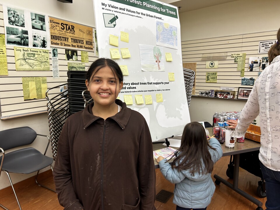 A young girl with light brown skin and dark brown hair tied back wearing a brought zip up jacket smiles in front of a large posterboard with sticky notes and children's drawings of trees in a well-lit room. 