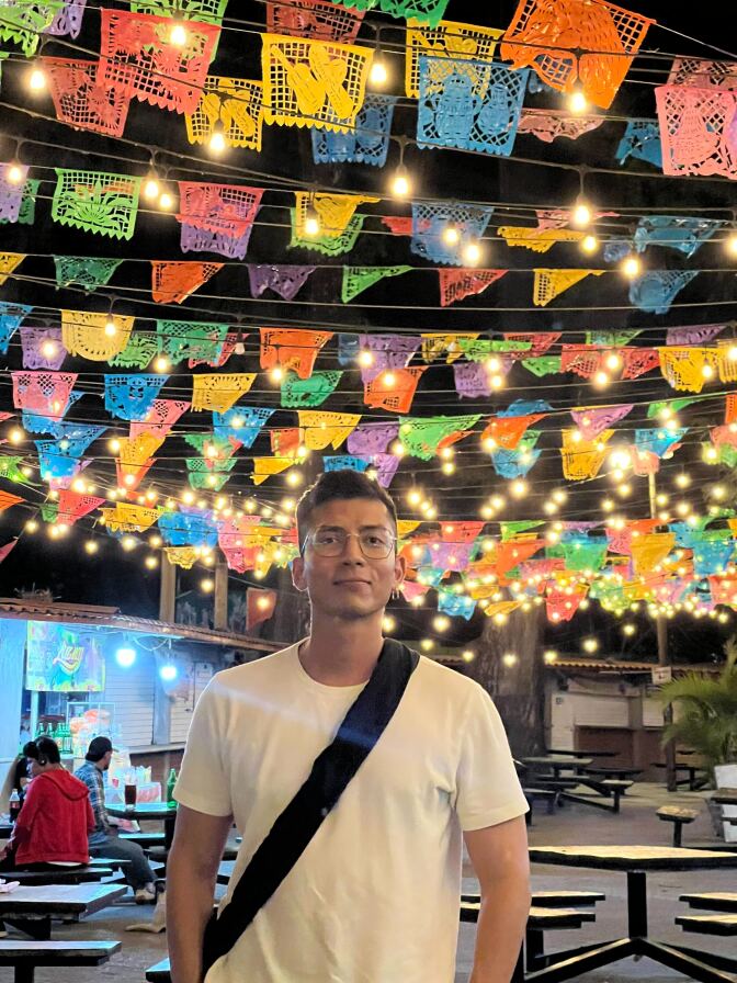 A brown-skinned Latino man stands in front of colorful flags. 