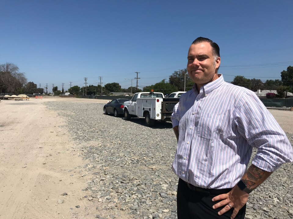 A man smiles at the camera with his hands on his hips standing in an empty lot.