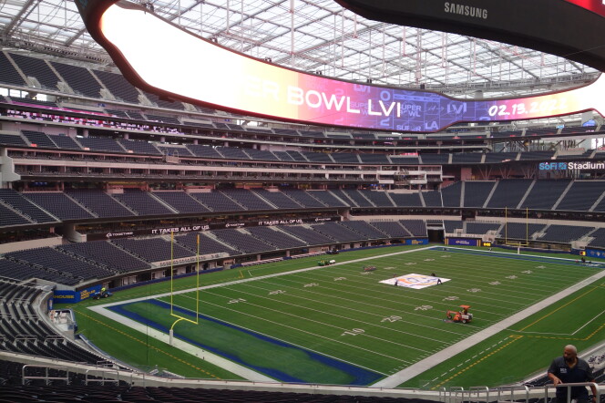 An interior view of the football field at SoFi Stadium in Inglewood. A large, ring-shaped video display board hangs from the roof of the stadium, displaying messages saying the stadium will host Super Bowl 56 on Feb. 13, 2022. Workers on seen preparing the field.