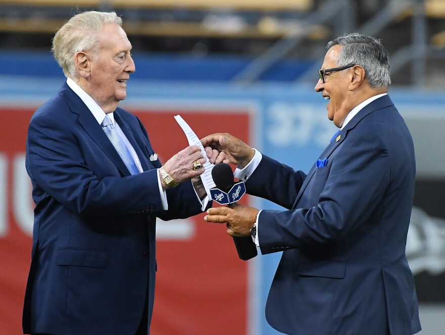 Vin Scully, wearing a navy suit and tie on the left, holds up a piece of paper. Jaime Jarrín, on the right, also dressed in a navy suit, holds a microphone in his left hand and reaches out toward the paper in Scully's hand. They appear to be outside at a baseball stadium, but the background is blurry.