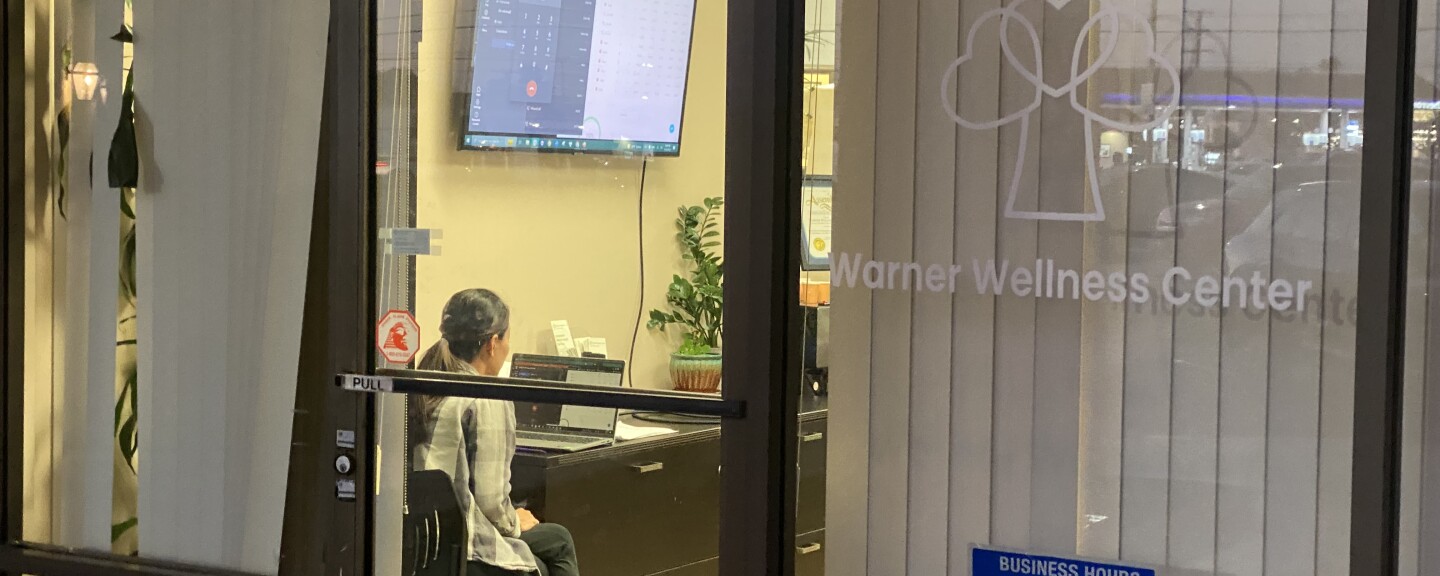 A person sits in an office with facing a TV screen showing a computer screen, with a logo by the door that says Warner Wellness Center