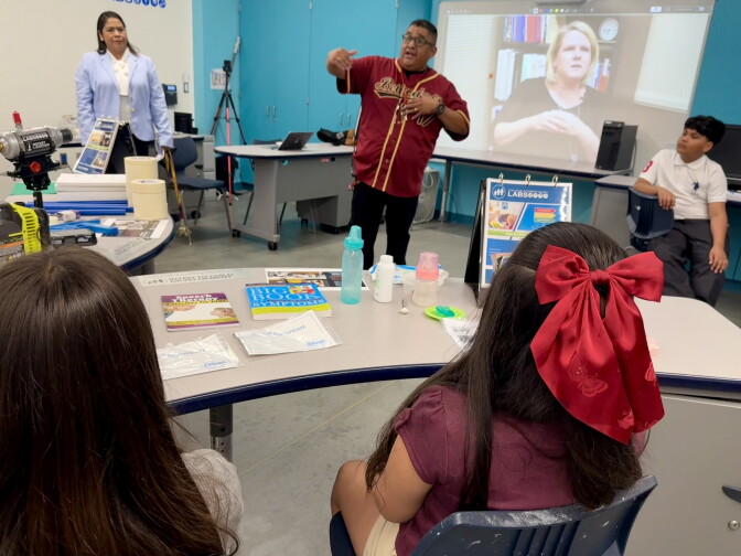 Students are seated and listening to a teacher, with medium skin tone and glasses, standing at the front of a classroom.