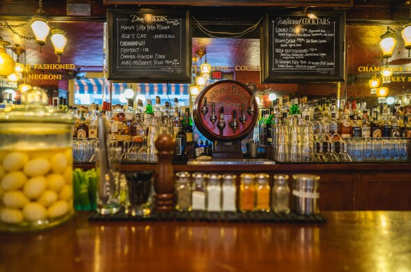 A warmly lit vintage bar interior at Cole’s, showcasing a gleaming back bar lined with glassware, colorful liquor bottles, and a signature “Littler Bros. Fountain” beer tap at center stage; a jar of pickled eggs and a neat row of spice shakers hint at the bar’s old-school charm and cocktail precision.