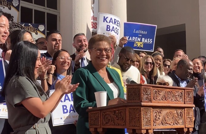Mayor-elect Karen Bass, dressed in a green pantsuit, gives remarks at a wooden podium as male and female supporters of different races applaud. 
