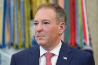 Man with light-toned skin, short brown and gray hair, looking off to the side. He is wearing a dark navy suit jacket over a white collared shirt, a red tie with a small repeating pattern, and a small American flag pin on the lapel.