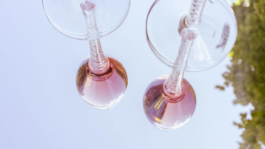 Two glasses of rosé wine are shown reflected on a glass table.