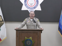 San Bernardino County Sheriff Shannon Dicus addresses a press conference from a podium. He wears a khaki uniform. 