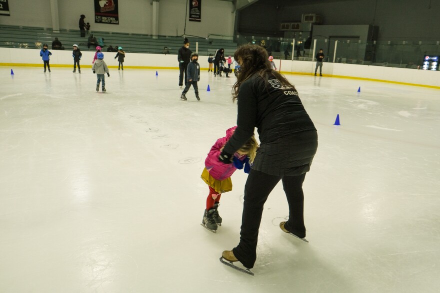 An adult dressed in black holds the hands of a young girl dressed in pink as they practice skating on an ice rink.