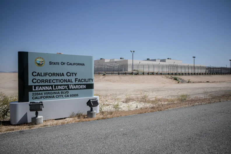 A outside shot of the California City Correctional Facility. The prison buildings are in the distance farther back and off to the right, and the sign for the prison is at the forefront and to the left of the frame.