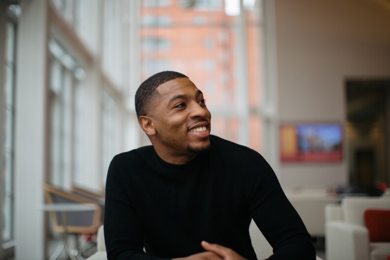 A man with medium dark skin tone wears a black long-sleeve shirt, smiles and looks toward his left shoulder.