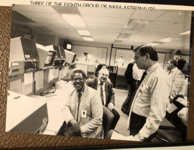A black and white photo of one of the rooms at the Apollo 6 launch. One light skinned man is smiling at the back, sitting down, looking at the camera. To his right is another light skinned man, who's looking at another dark skinned man sitting down in a chair. That man is  also smiling and looking at the camera. They are all wearing suits and ties.