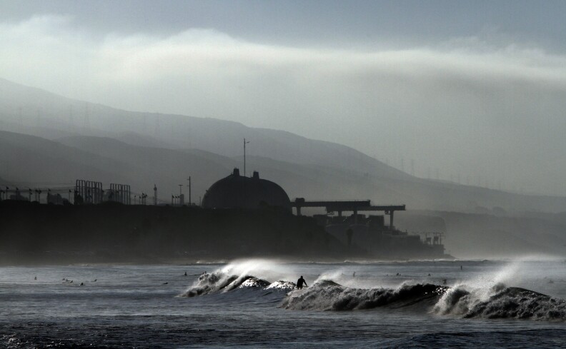 Surfers float in the water and catch waves. White mist rises from the breaking waves. In the background are two large domed buildings and industrial infrastructure.