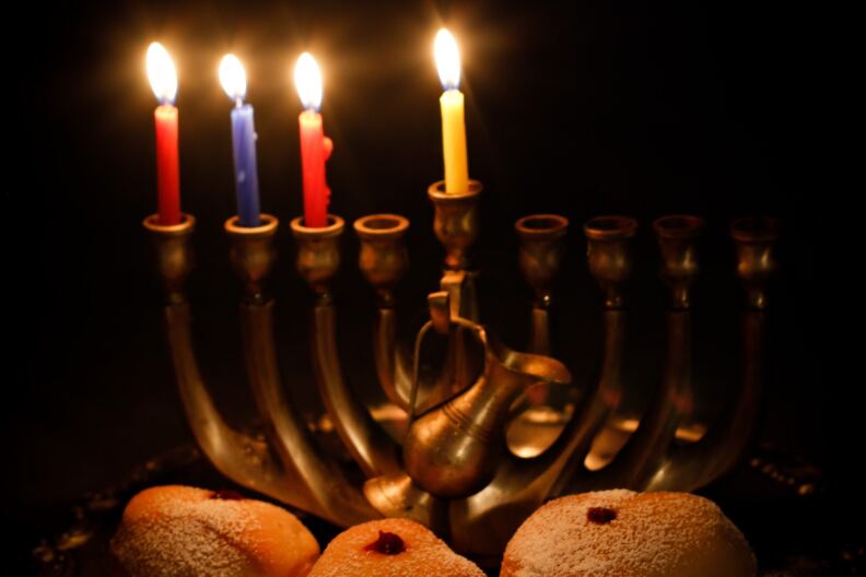 A menorah with four lit candles sits above jelly donuts and dreidels.