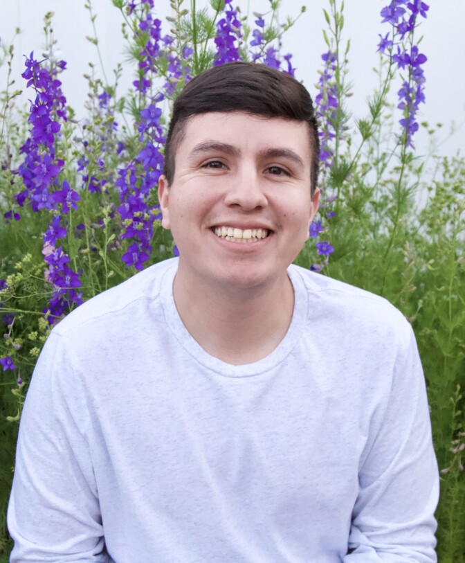 Ivan Beas wears a light-colored long-sleeve T-shirt. Beas has short brown hair and poses outside in from of purple flowers.