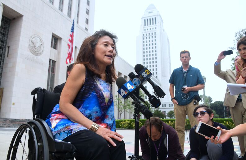 A woman in a wheelchair speaks into mics with TV-station flags as reporters record her. L.A. City Hall is in the background.