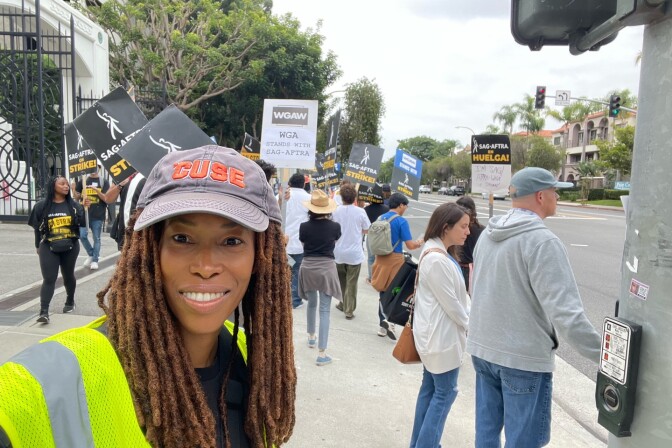 Actor Renée Threatte looks towards the camera smiling. She wears a baseball style cap and a yellow safety vest. Picketing writers and actors can be seen on the sidewalk behind her. 