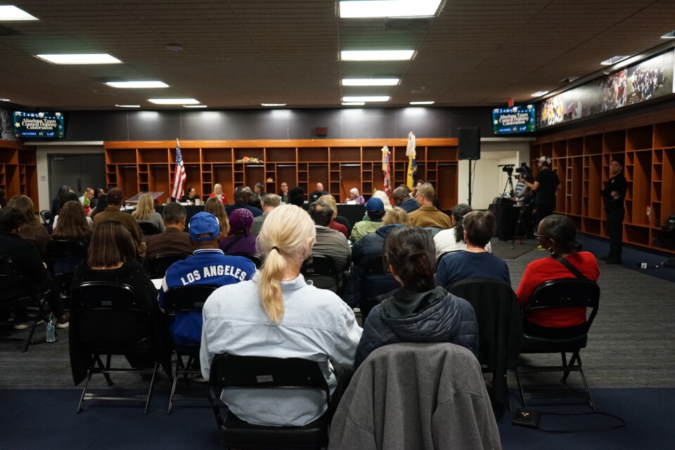 A sports locker room is filled with people seated in folding chairs. They're facing away from the camera and towards a panel. There is a cameraperson on the right.