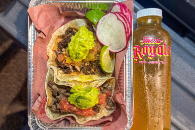 Two carne asada tacos topped with guacamole, salsa and diced onion in a foil tray lined with brown paper, garnished with sliced radish and lime wedges, beside a clear bottle of amber jasmine tea labeled "Tacos Royale."