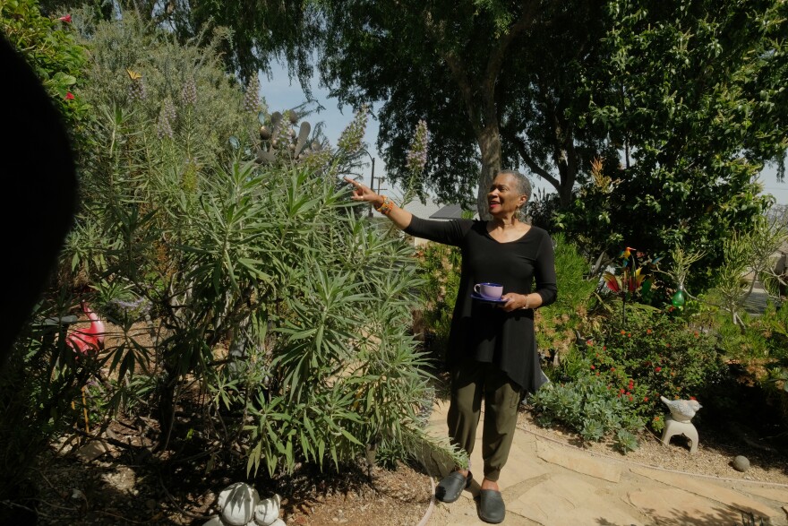 A woman wearing a black shirt and green pants points to a butterfly alighting on a large plant in her front yard. Trees and other plants rise up in the background.