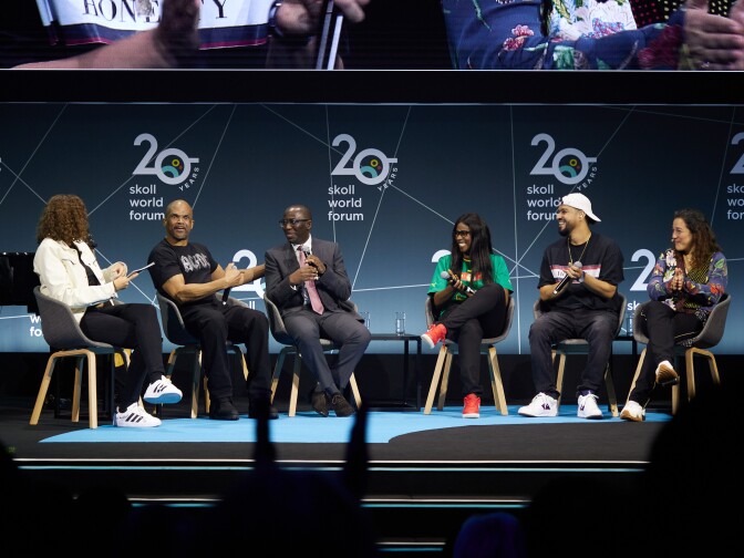A panel of six men and women are sitting in chairs and talking on an illuminated stage. 