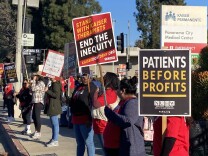 Striking mental health workers stand in line along a street in Panorama City. They hold signs that read "Patients before profits" and "End the inequity" 