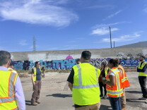 People in high-visibility safety vests stand near a graffiti-covered concrete wall outdoors.