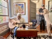 An elderly man with white hair plays guitar, while a large goldendoodle dog jumps on a hispanic older man wearing a hat. 