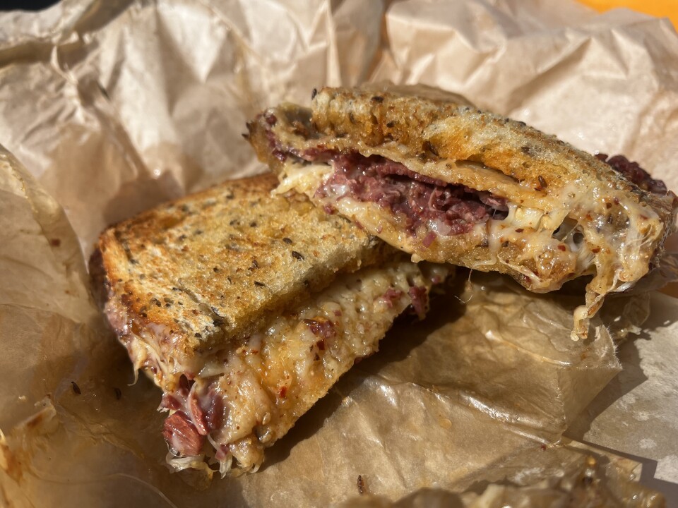 A sandwich is cut into two halves, with one half sitting partially on top of the other on a light brown to-go wrapper. The bread is toasted to a light golden brown color, with dark flecks embedded throughout. The contents of the sandwich include melted cheese along with shreds of cooked meat that are dark reddish-pink in color.