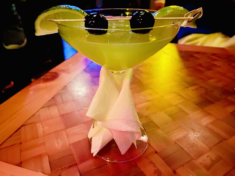 A martini glassis filled with a bright green liquid, topped with two dark berries speared and positioned to look like eyes, and two .lime wedges placed on the rim of the glass to look like ears. The result looks like Grogu, aka Baby Yoda.