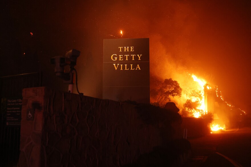 A wildfire is burning trees and structures behind a large black and gold sign that reads "The Getty Villa"