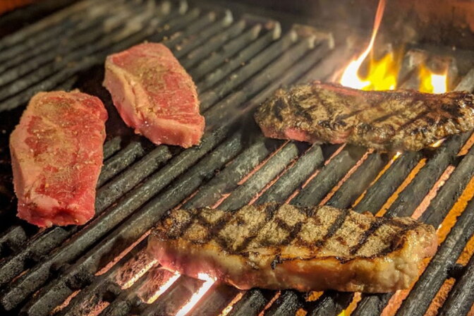 Four raw and grilled steaks cooking on a flaming charcoal grill, two seared with grill marks and two still raw.