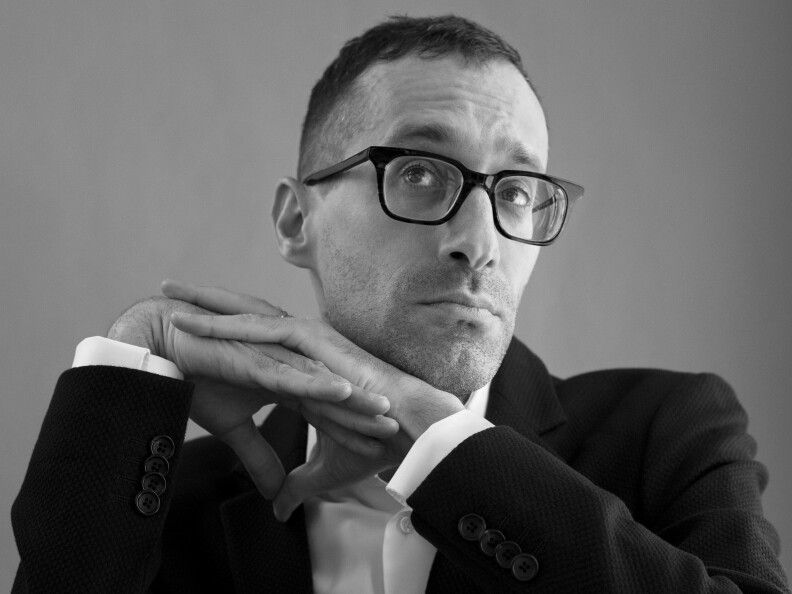 A black and white photo shows a man with glasses, short-cropped hair and dressed in a white business shirt and black jacket, looking up and to the left, hands clasped at an angle to the right of his face.