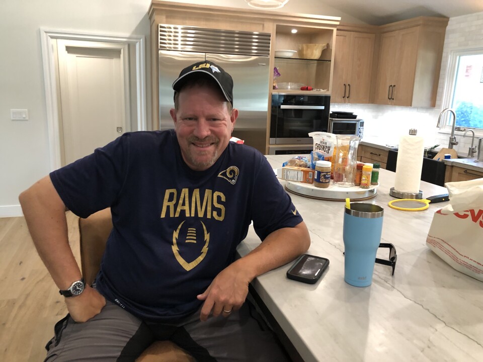 A man sits on a kitchen stool, his left elbow leaning on a kitchen island. He's wearing a shirt and hat that both support the Los Angeles Rams.