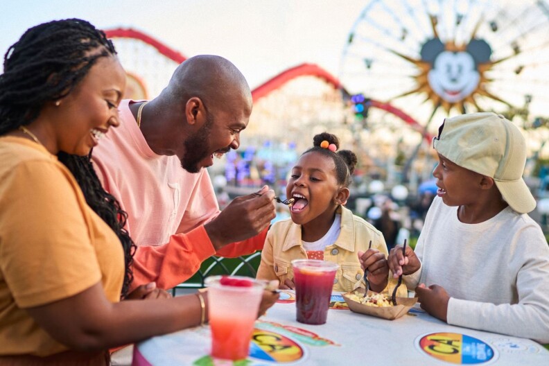 A photo of a family of four, mom, dad and two kids, eating in front of the Mickey Mouse-themed ferris wheel at California Adventure. The father is feeding his daughter.